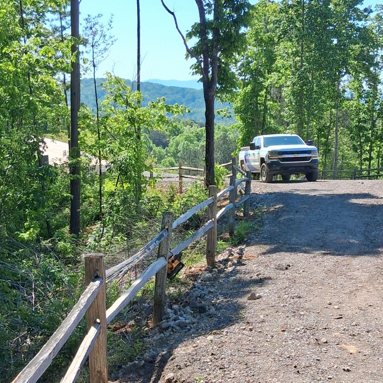 White pickup truck on a gravel forest road beside a wooden fence, with mountains in the background