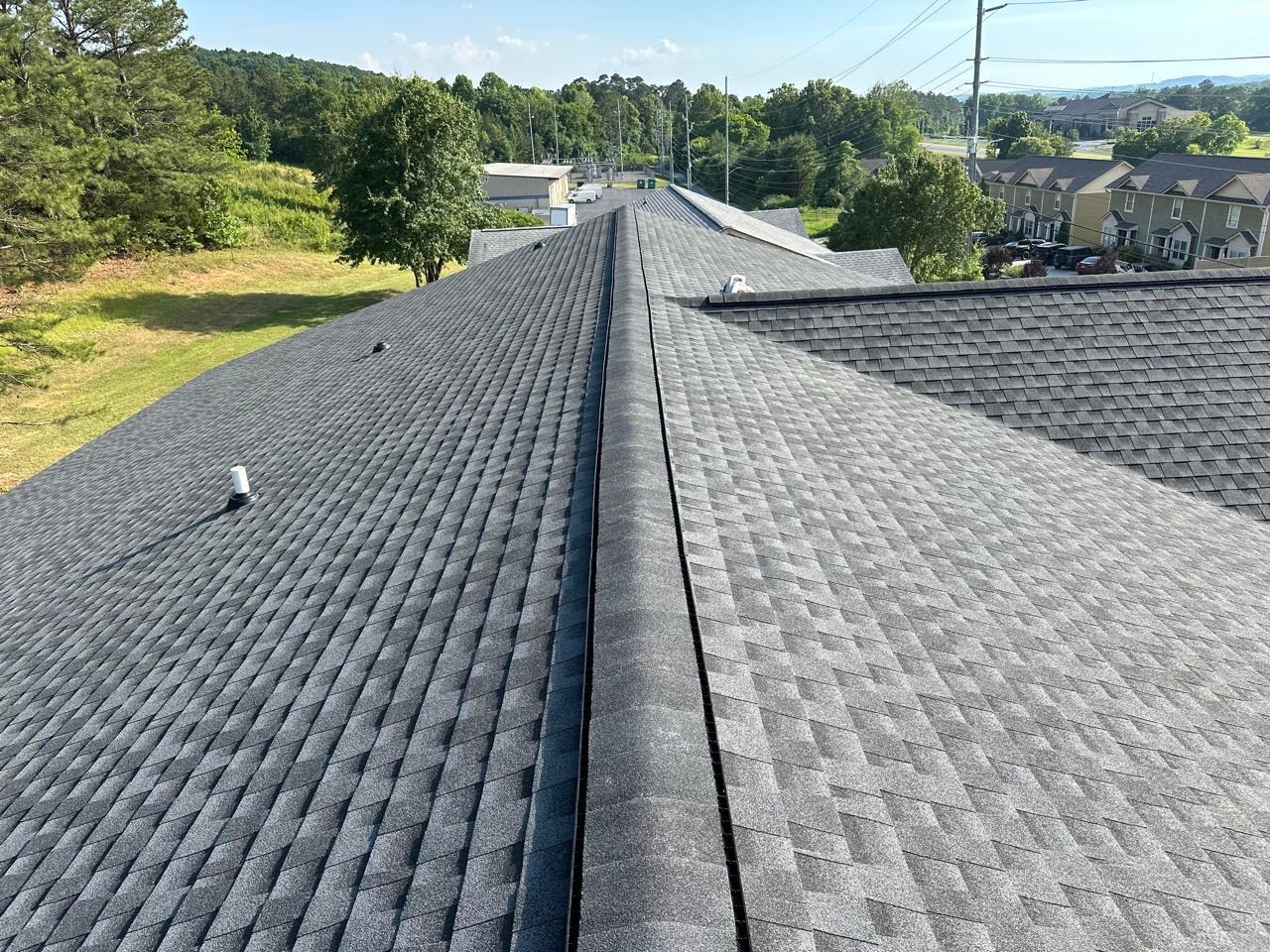 Two gray shingled roofs meeting in a valley, with trees and homes in the background under a clear sky
