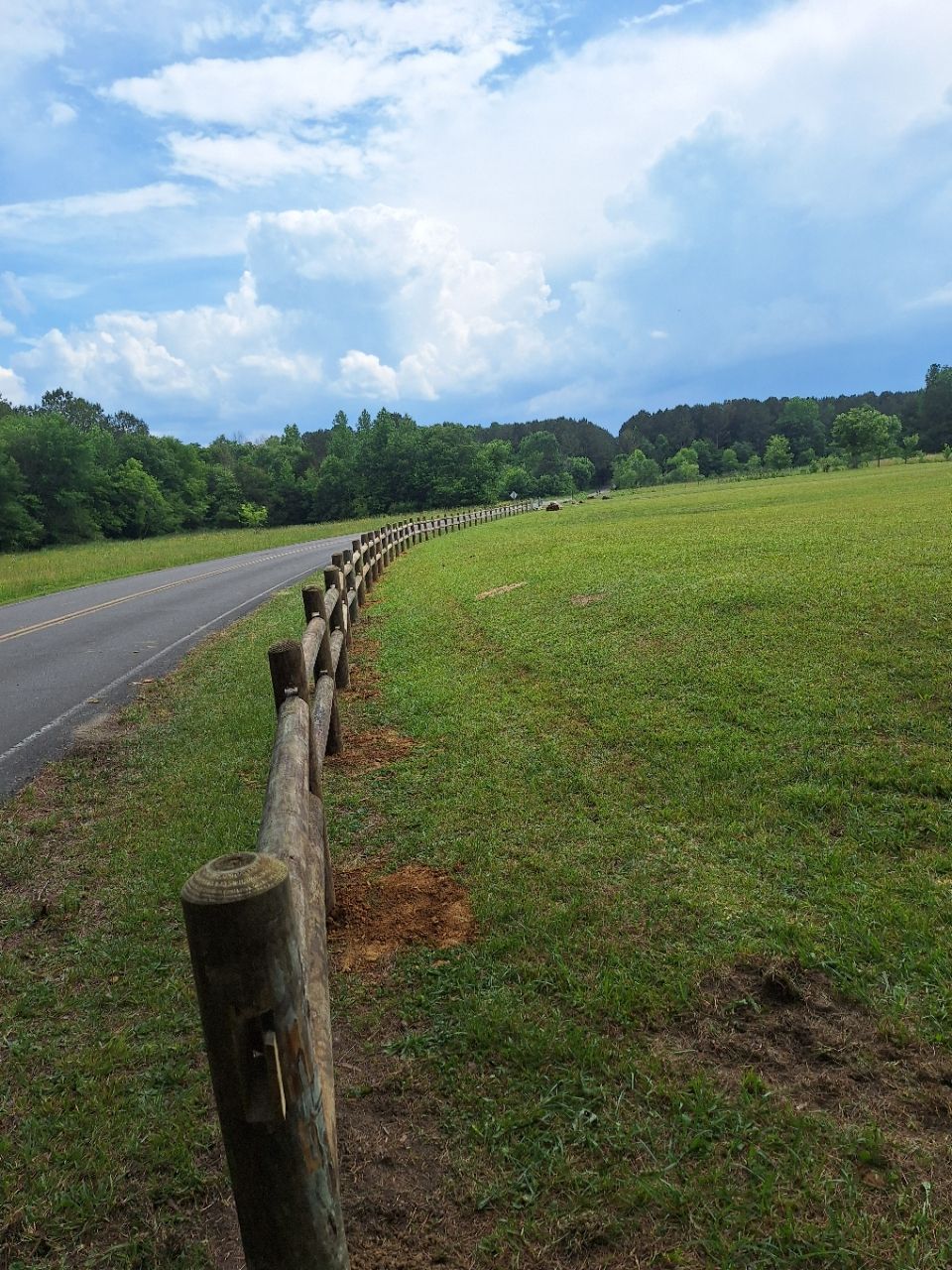 Curved wooden fence beside a paved road through a green field under a cloudy sky