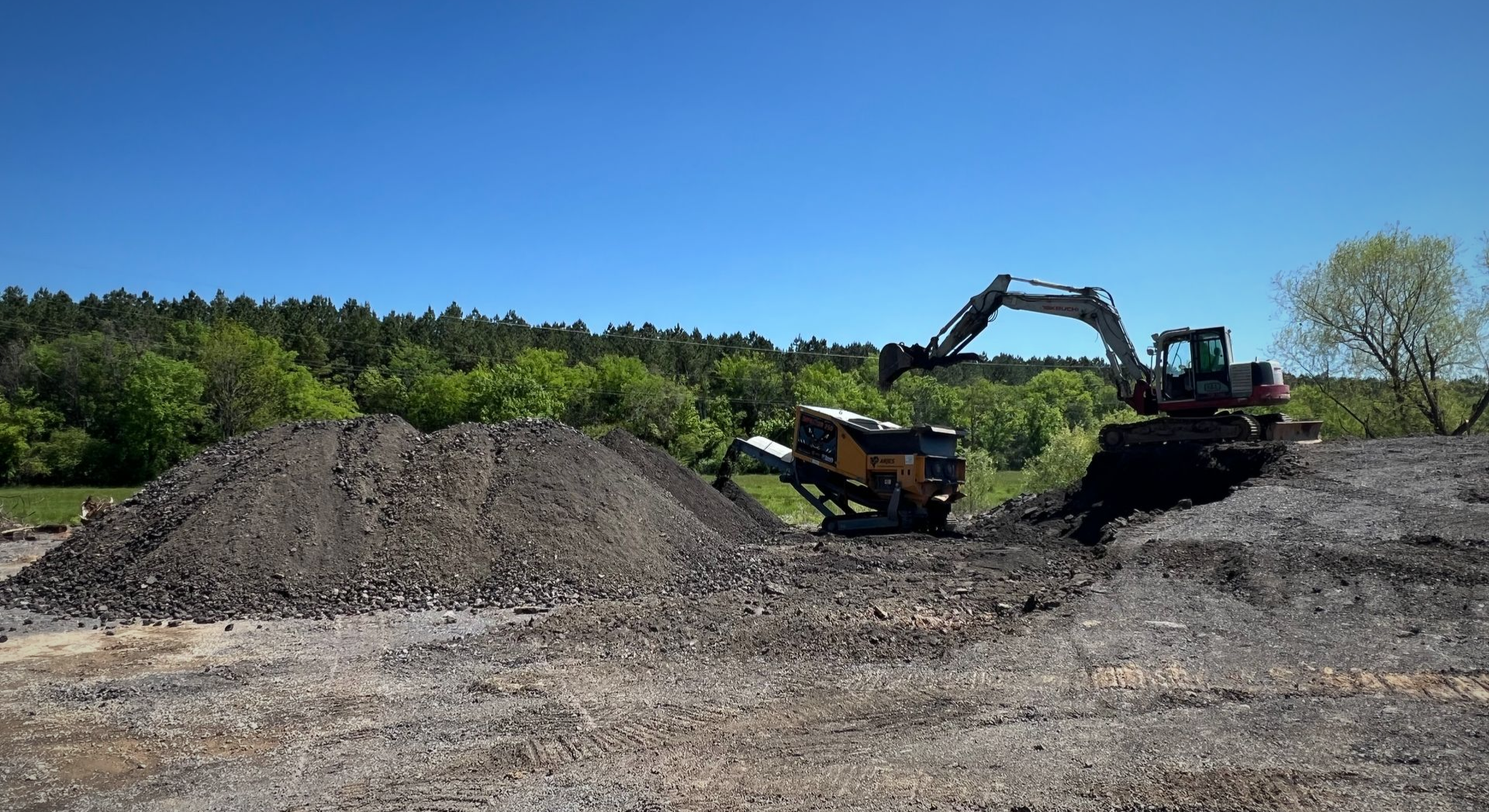 Two excavators digging dirt beside a large gravel mound on a sunny construction site