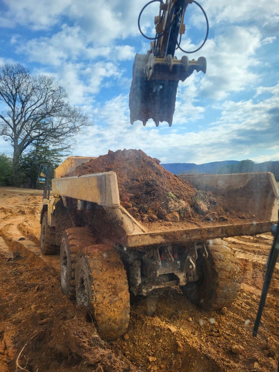 Excavator loading soil into a dump truck at a muddy construction site under a cloudy blue sky