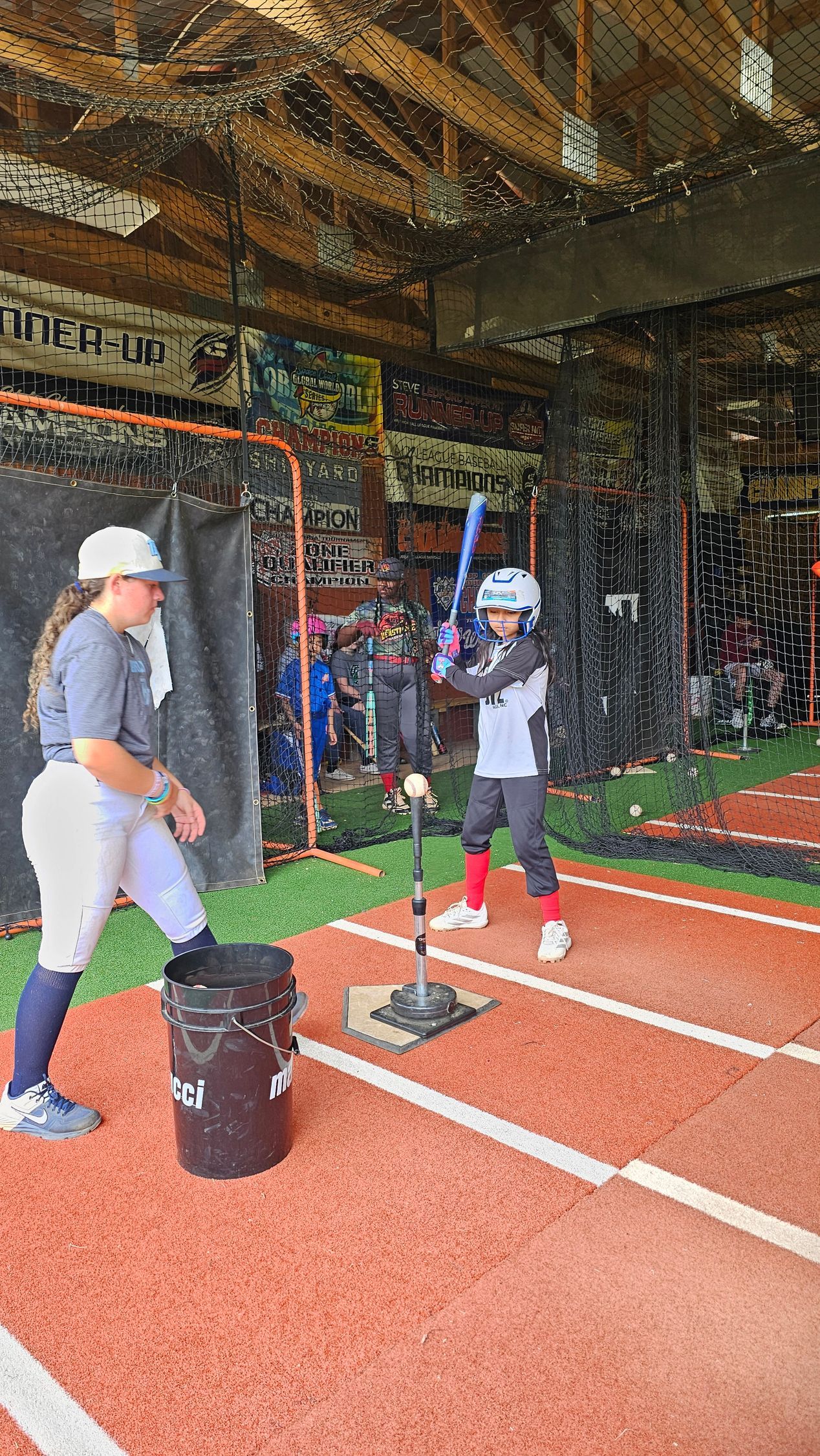 Two people in a batting cage, one swinging at a tee on an indoor turf field.