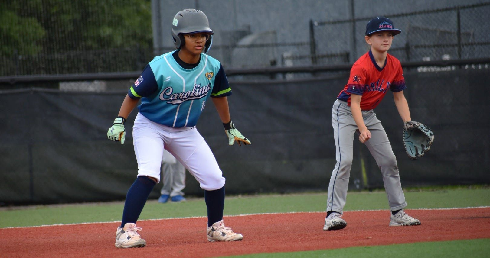 Two baseball players in helmets poised on base, on an infield near a fence.