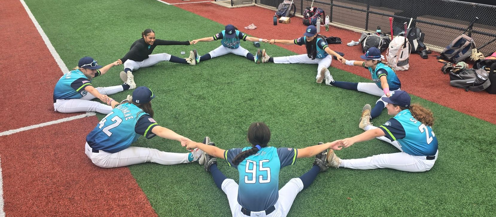 Baseball players in teal jerseys stretching in a circle on turf near the track and dugout area.
