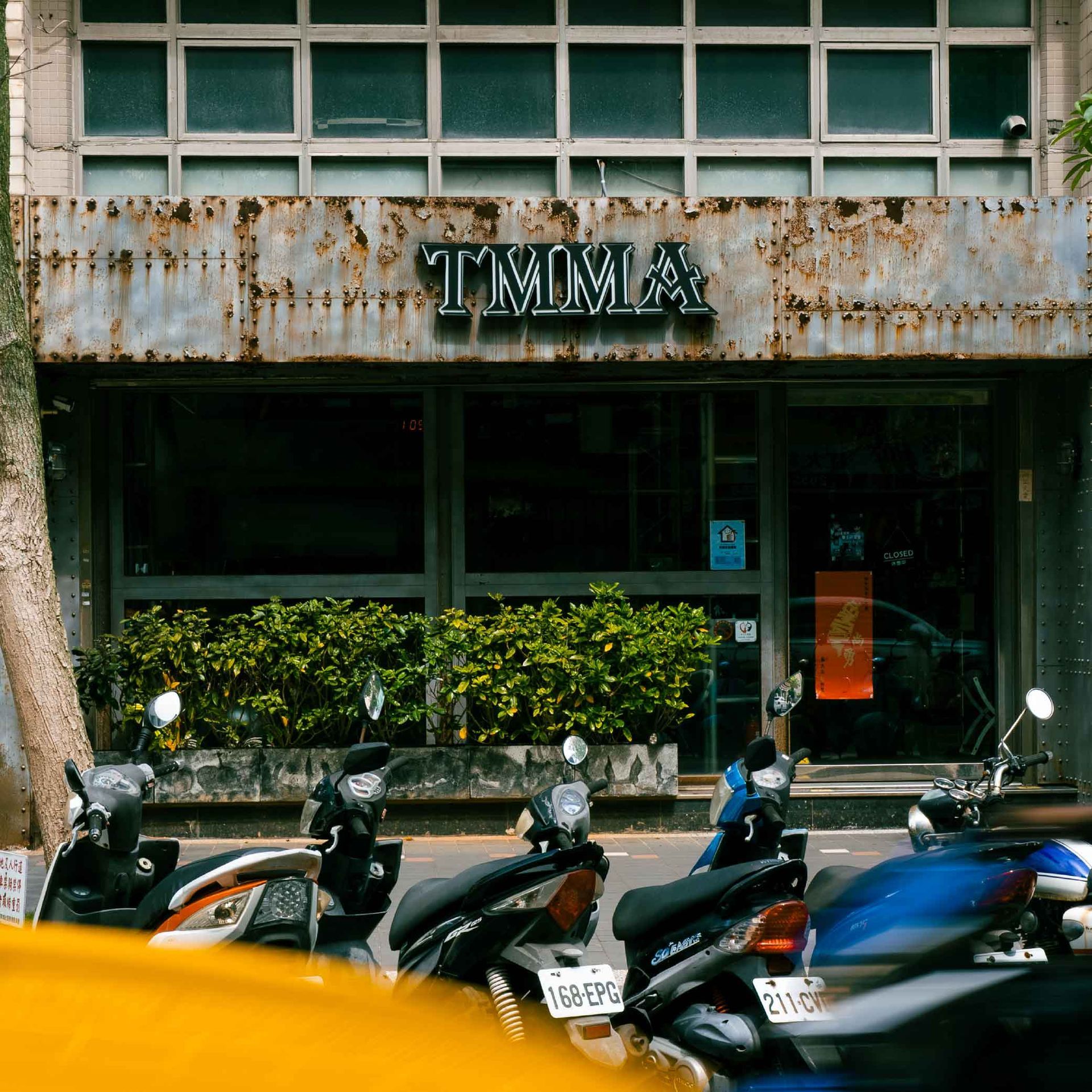 金屬工業風的店面掛著TMMA的招牌，幾輛摩托車停在前方，一輛黃色汽車經過形成殘影。