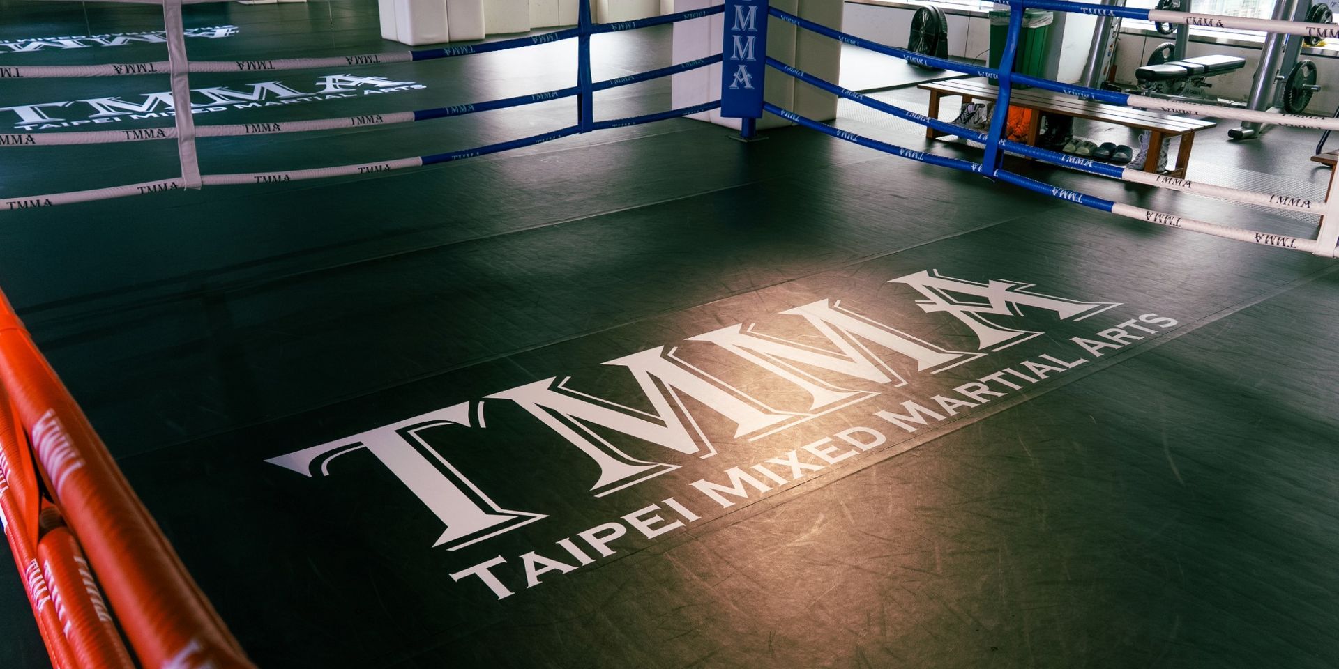 空曠的拳館內，架設一座繩圈擂台，地墊上印有 TMMA Taipei Mixed Martial Arts 標誌字樣。