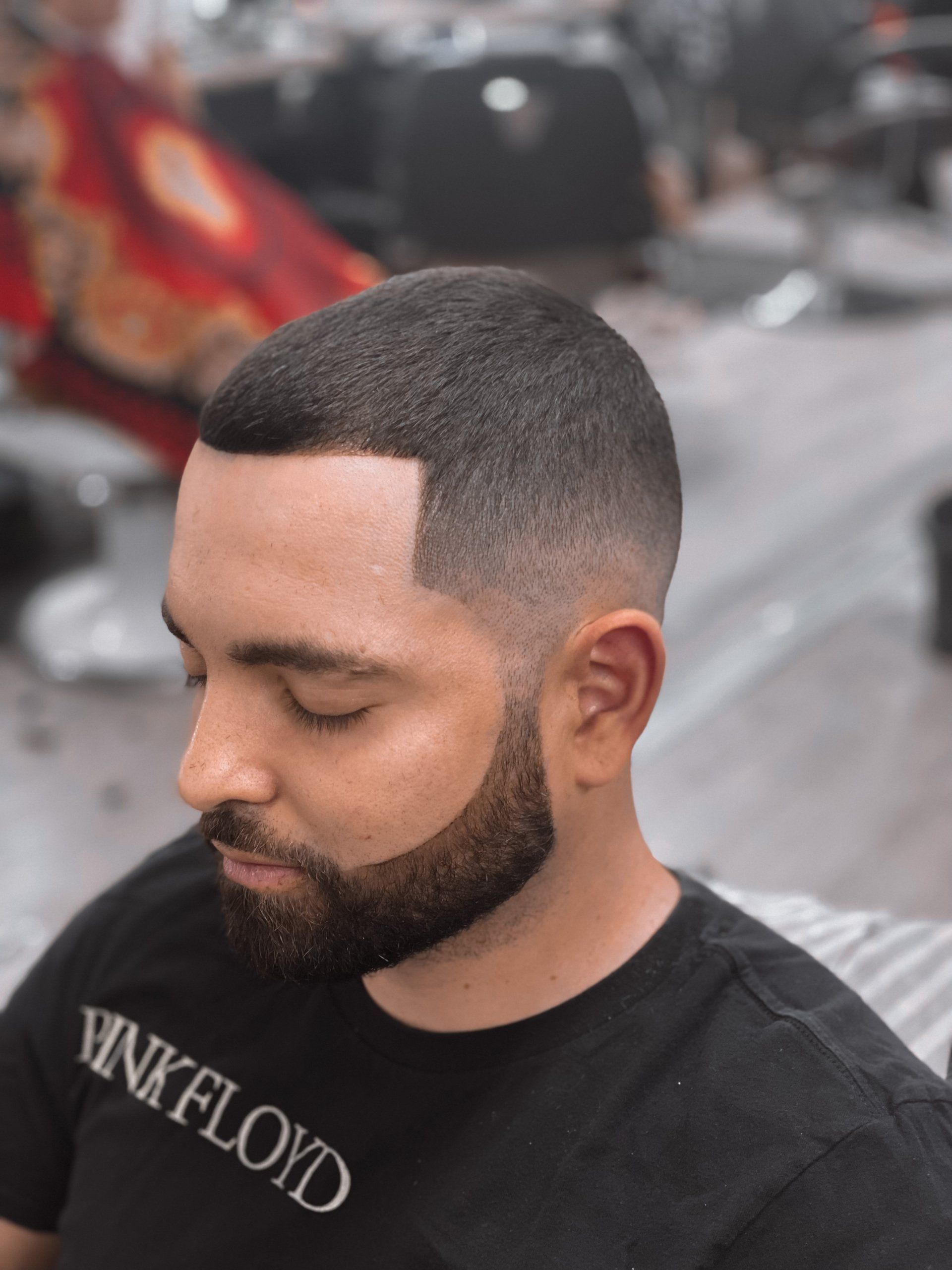 Close-up of a man with a sharp fade haircut and beard, looking down in a barbershop chair.