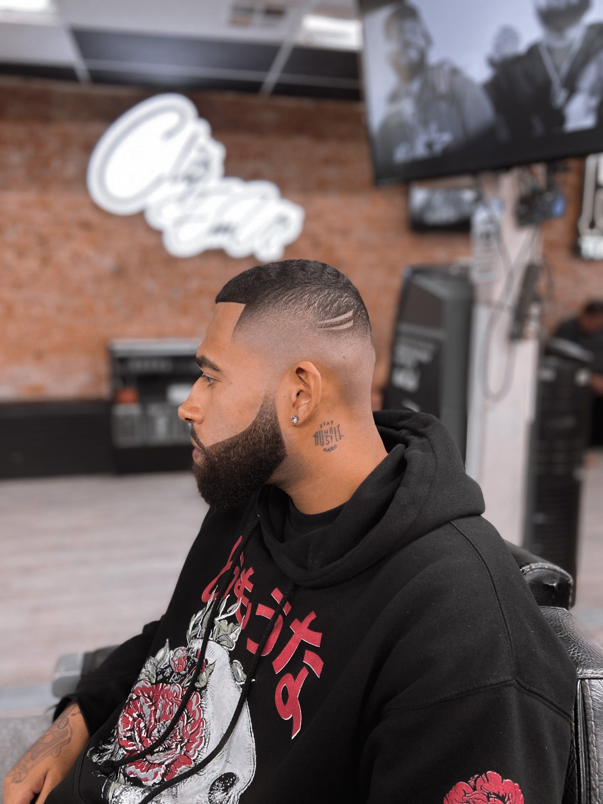 Man in black hoodie seated in a barbershop, shown in profile against a brick wall.