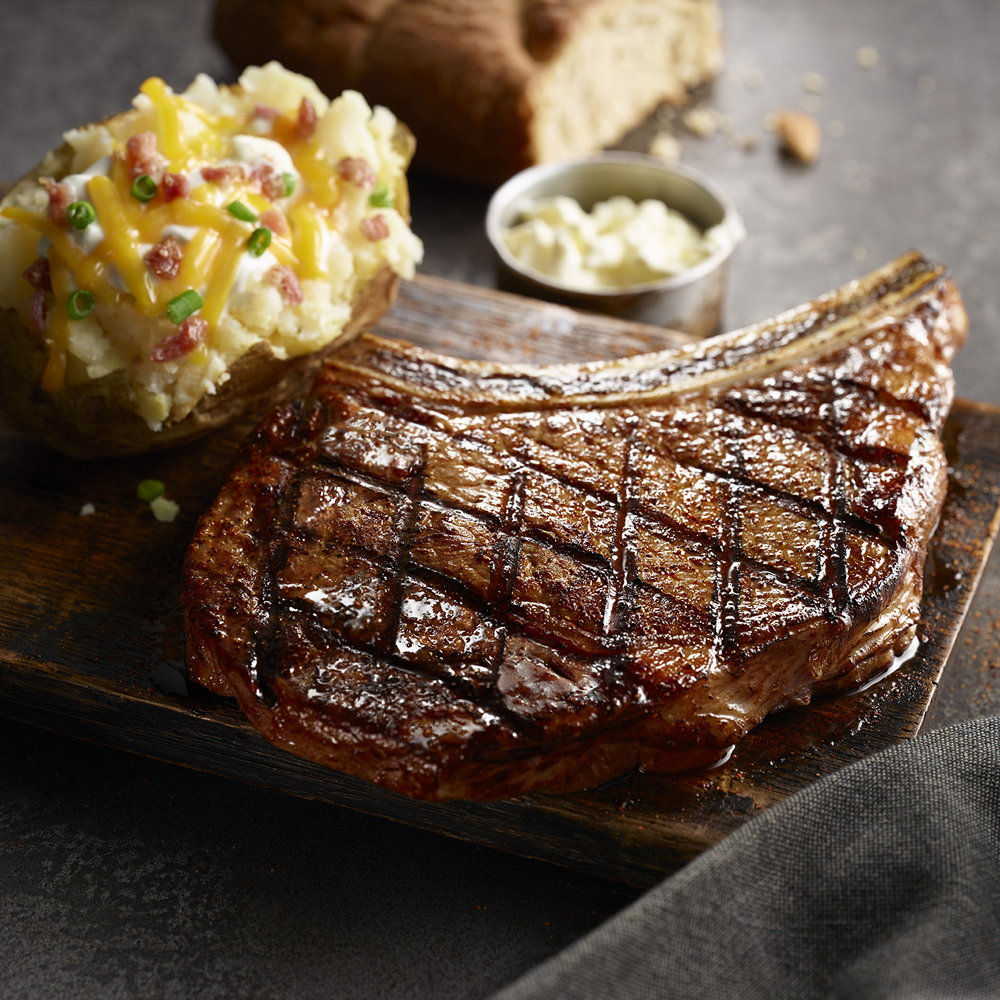A steak with a baked potato and mashed potatoes on a wooden cutting board.