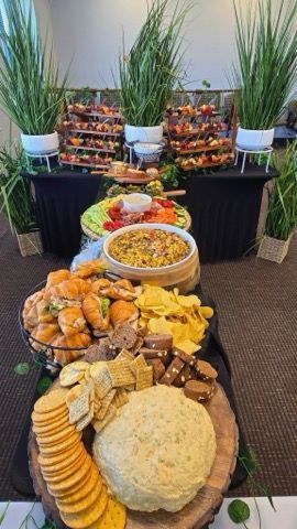 A catered food display featuring dips, crackers, sandwiches, chips, and appetizers. Green plants decorate the space.
