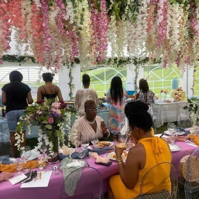 People at an outdoor event with hanging flower decor, seated at tables with food and drinks.