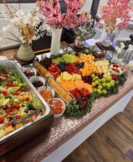 Buffet spread with salad, fruit, cheese, crackers, and flower arrangements on a rose gold tablecloth.
