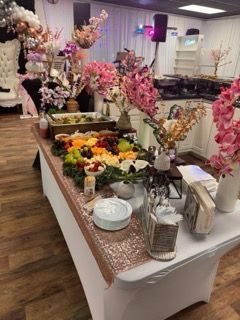 Buffet table with fruit, flowers, and food at a decorated event with a white backdrop.