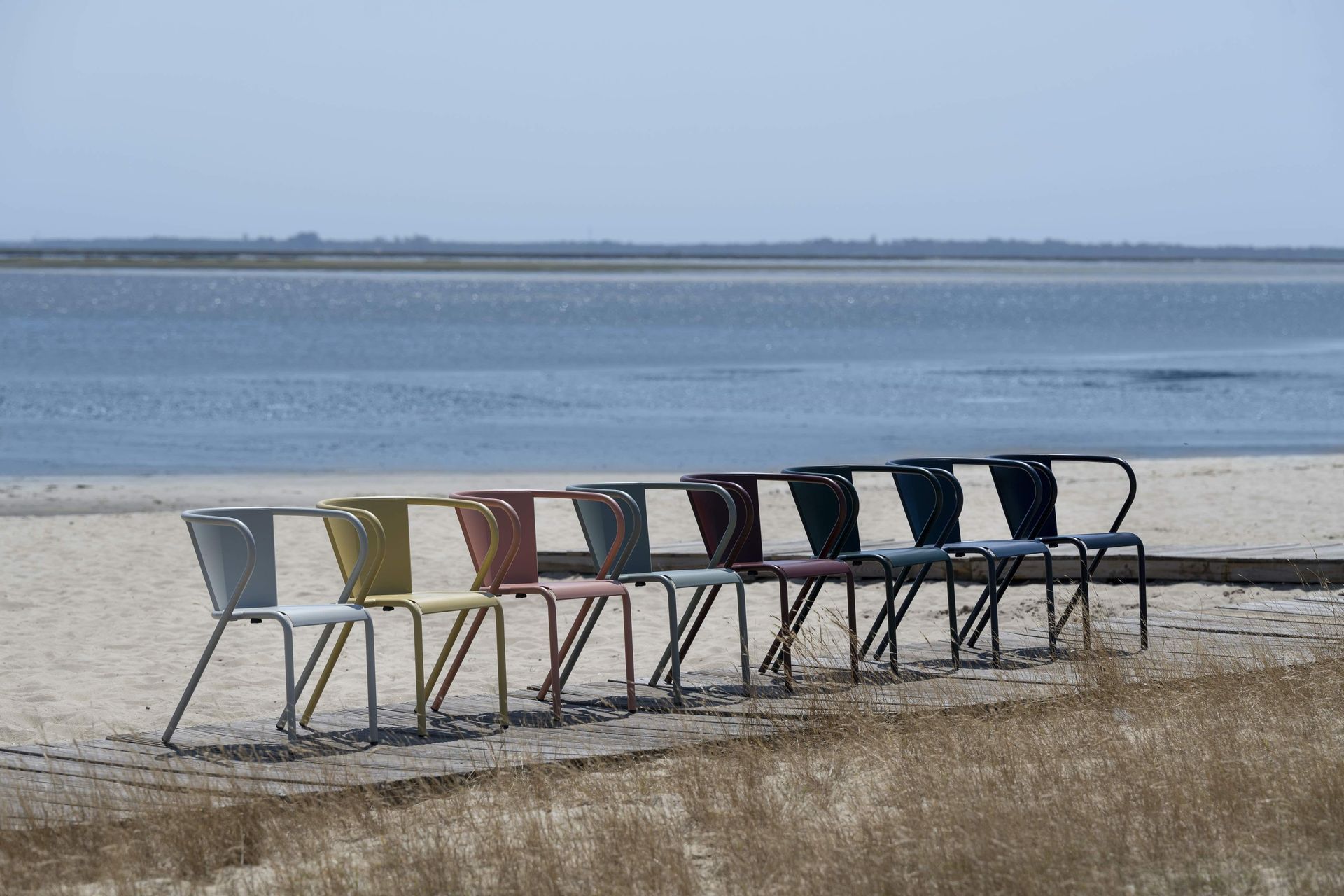 Een rij van acht kleurrijke metalen stoelen staat opgesteld op een zandstrand, tegenover een kalm wateroppervlak.