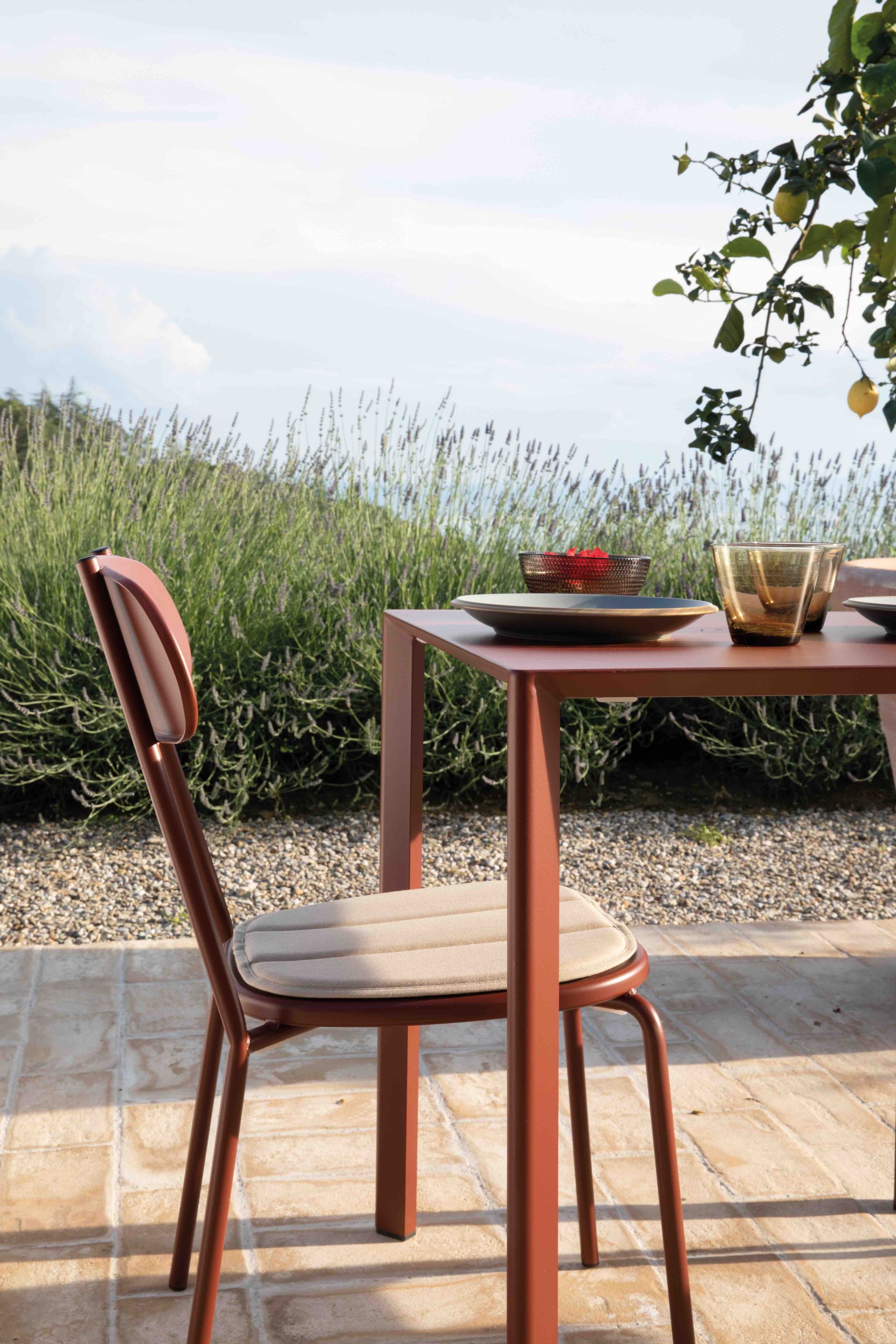 Een bruine metalen stoel met een beige kussen staat bij een bijpassende tafel op een zonnig terras, vlakbij citroenbomen en -struiken.