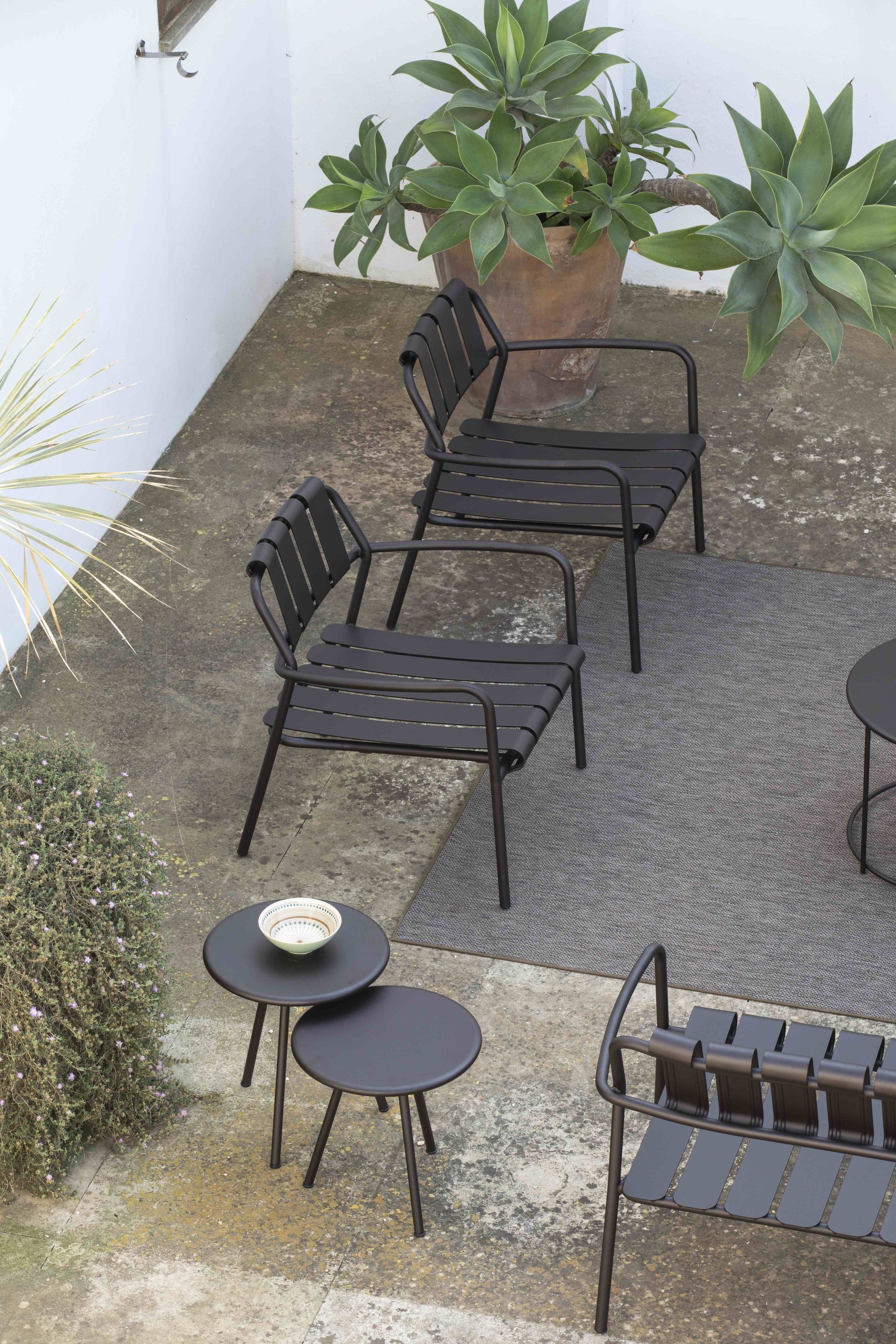 Een terras met donkergrijze stoelen, kleine ronde bijzettafels en grote vetplanten in potten op een stenen vloer.