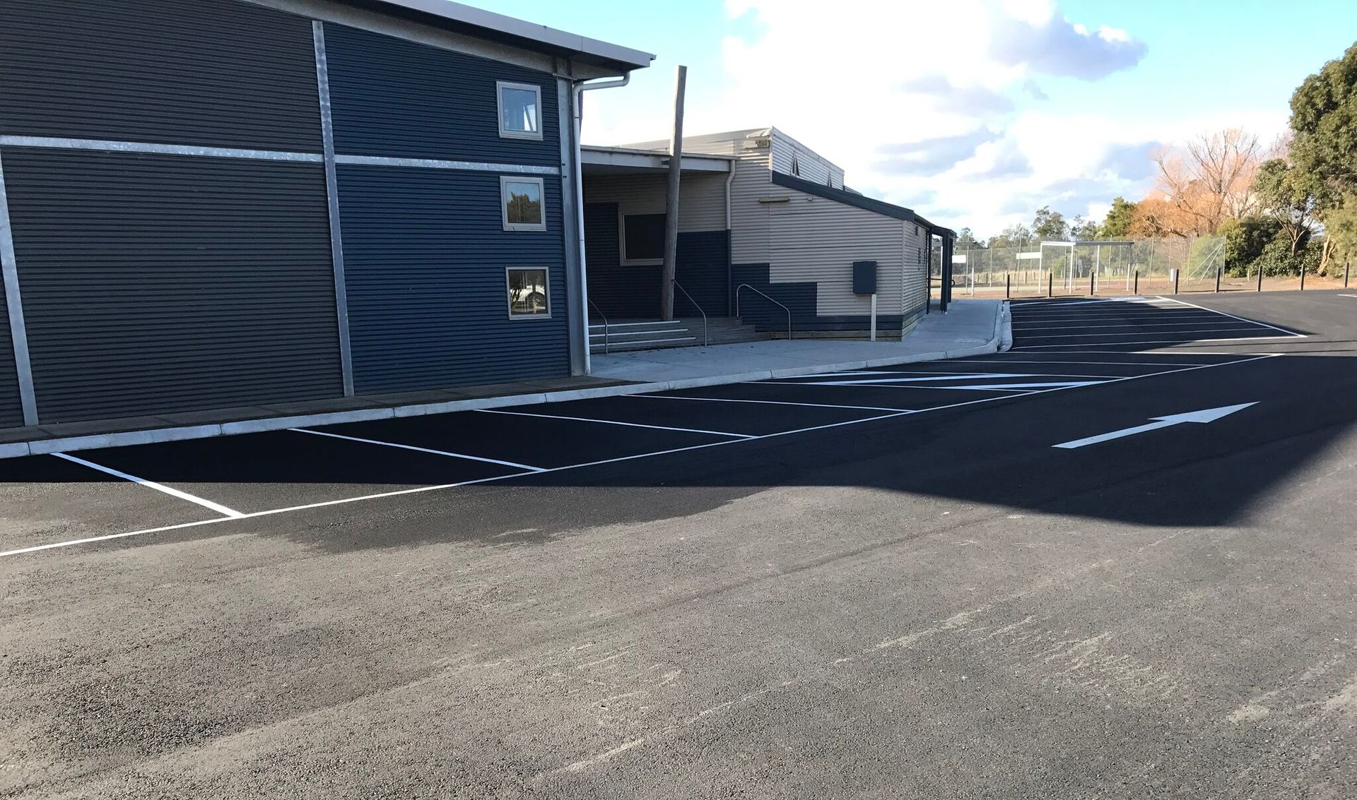 Empty parking lot beside a dark building on a sunny day, with painted lines and long shadows.