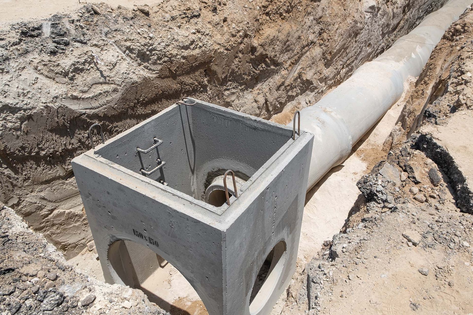 Concrete utility box beside a large buried pipe in an excavated dirt trench