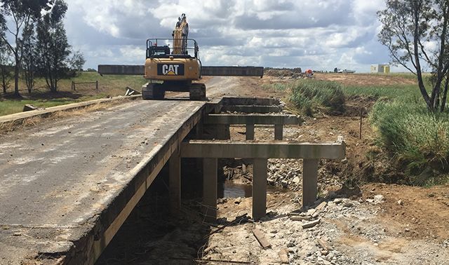 Excavator on a damaged road bridge over a dry ditch in a rural area under cloudy skies