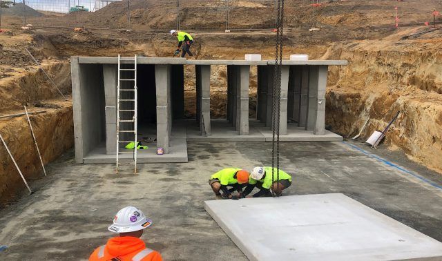 Construction workers on a concrete underground structure with excavation around it, one on the roof and one on a ladder.