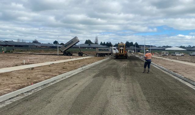 Construction crew paving a new road under a cloudy sky, with machinery and workers visible along the roadway.