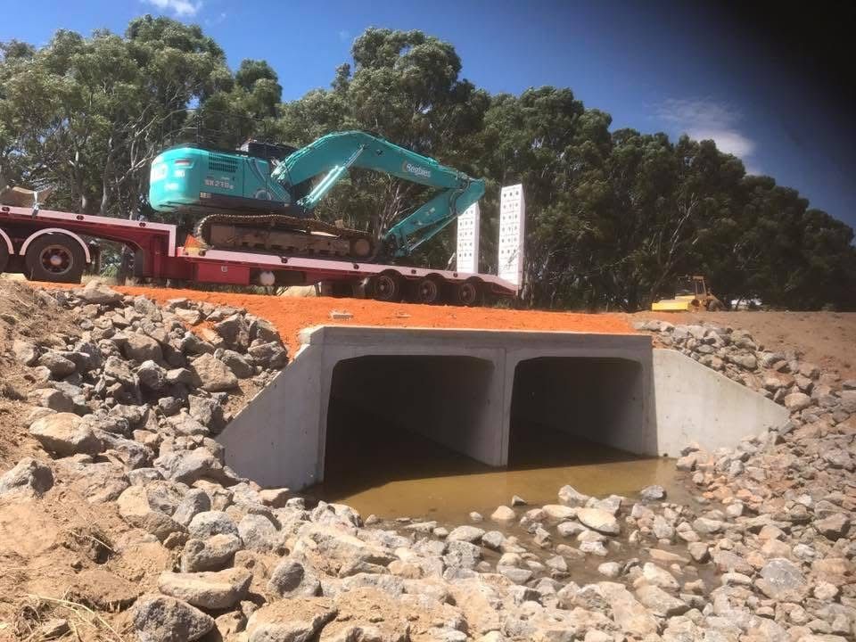 Construction truck unloading a teal excavator into a concrete culvert beside a rocky roadside