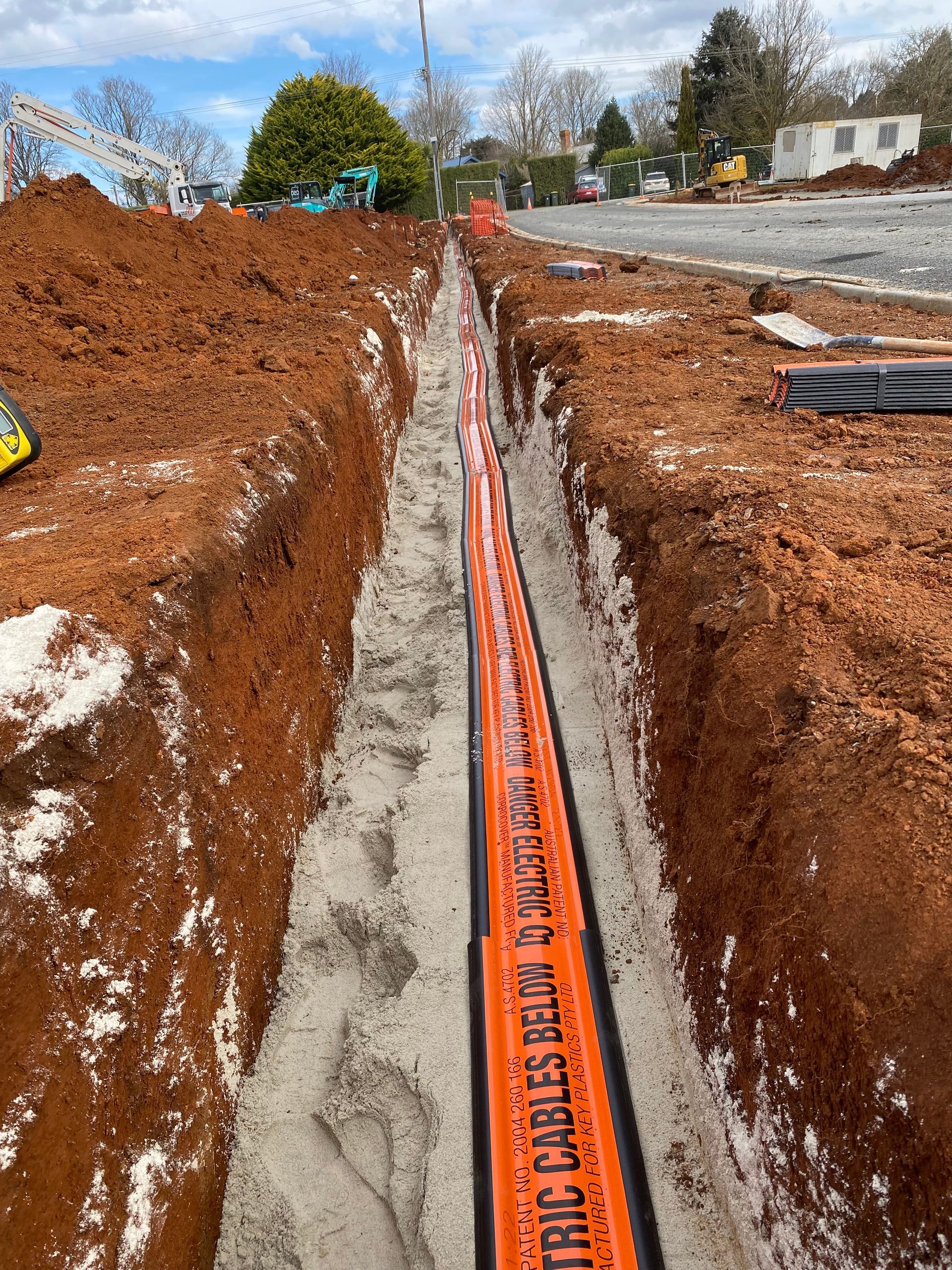 Orange utility conduit in a narrow trench at a construction site, with dirt piles and houses in the background