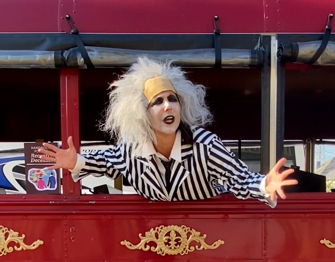 Person with wild white hair leans from a red tram, arms outstretched, wearing a black-and-white striped jacket.