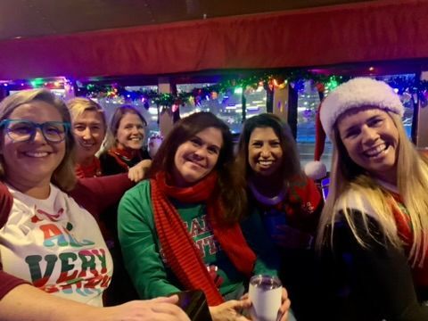Five friends smiling in festive holiday sweaters and hats at an indoor party with colorful lights