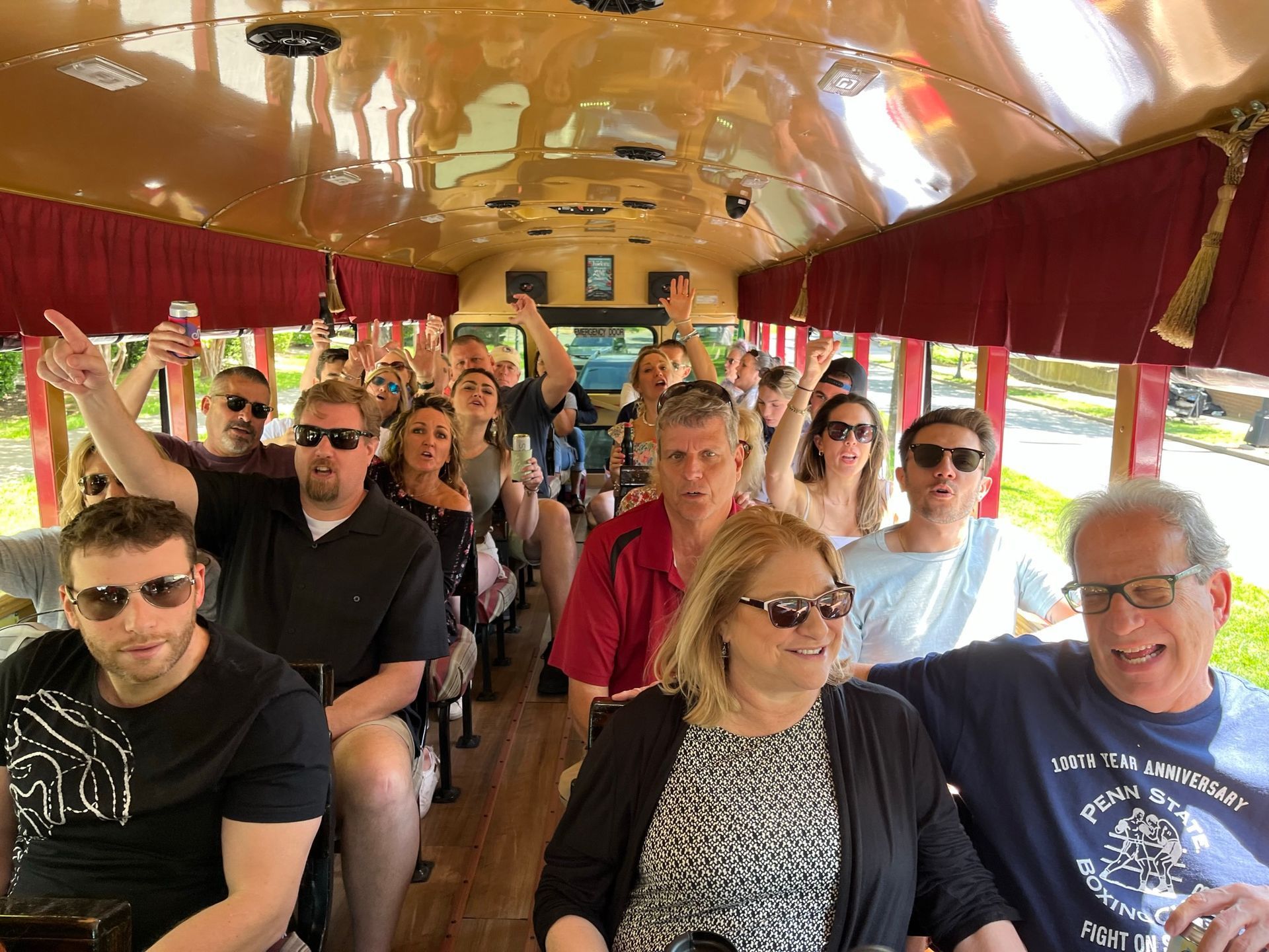Passengers sitting inside a red-canopy open tour tram, many smiling and raising hands.