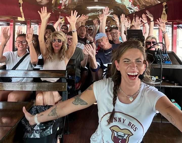 Friends smiling and waving on a bus tour, with a woman in front grinning at the camera.