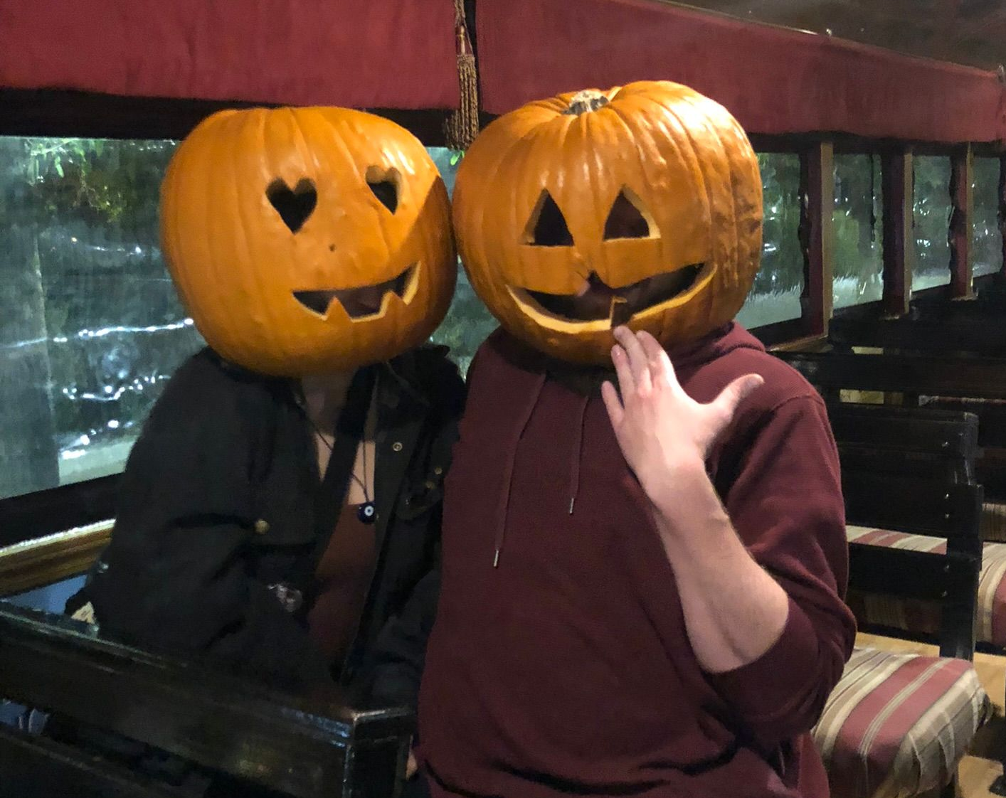 Two people with pumpkin jack-o'-lantern masks sitting on a train bench, one waving.