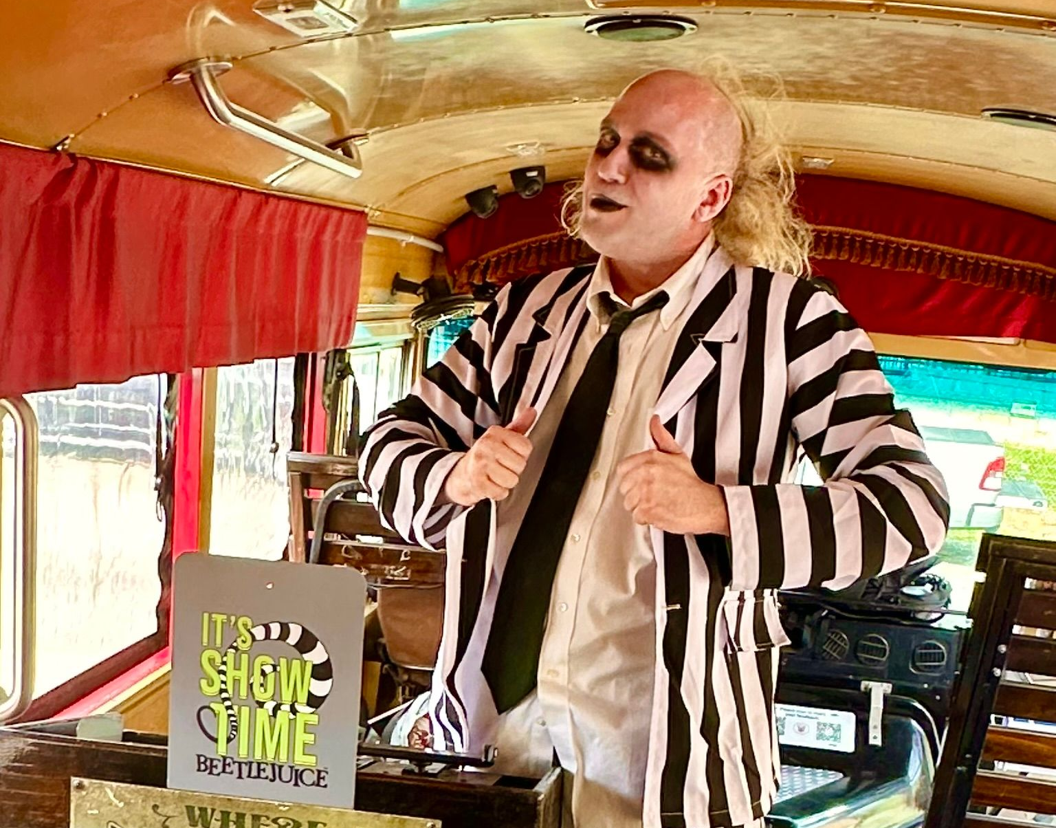 Man in a black-and-white striped jacket singing on a bus, with red curtains and seats around him.