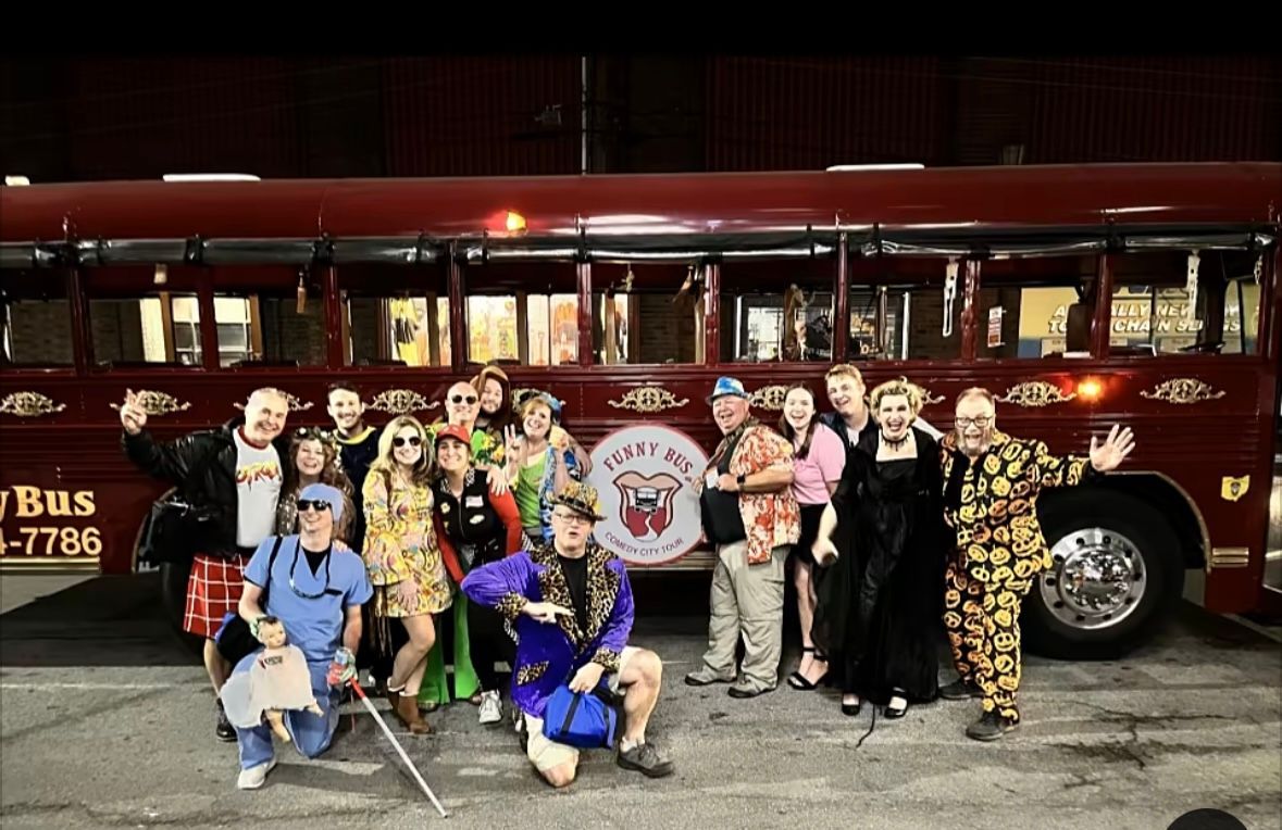 Group posing in front of a maroon bus at night, with one person holding a colorful bouquet and others in costumes.