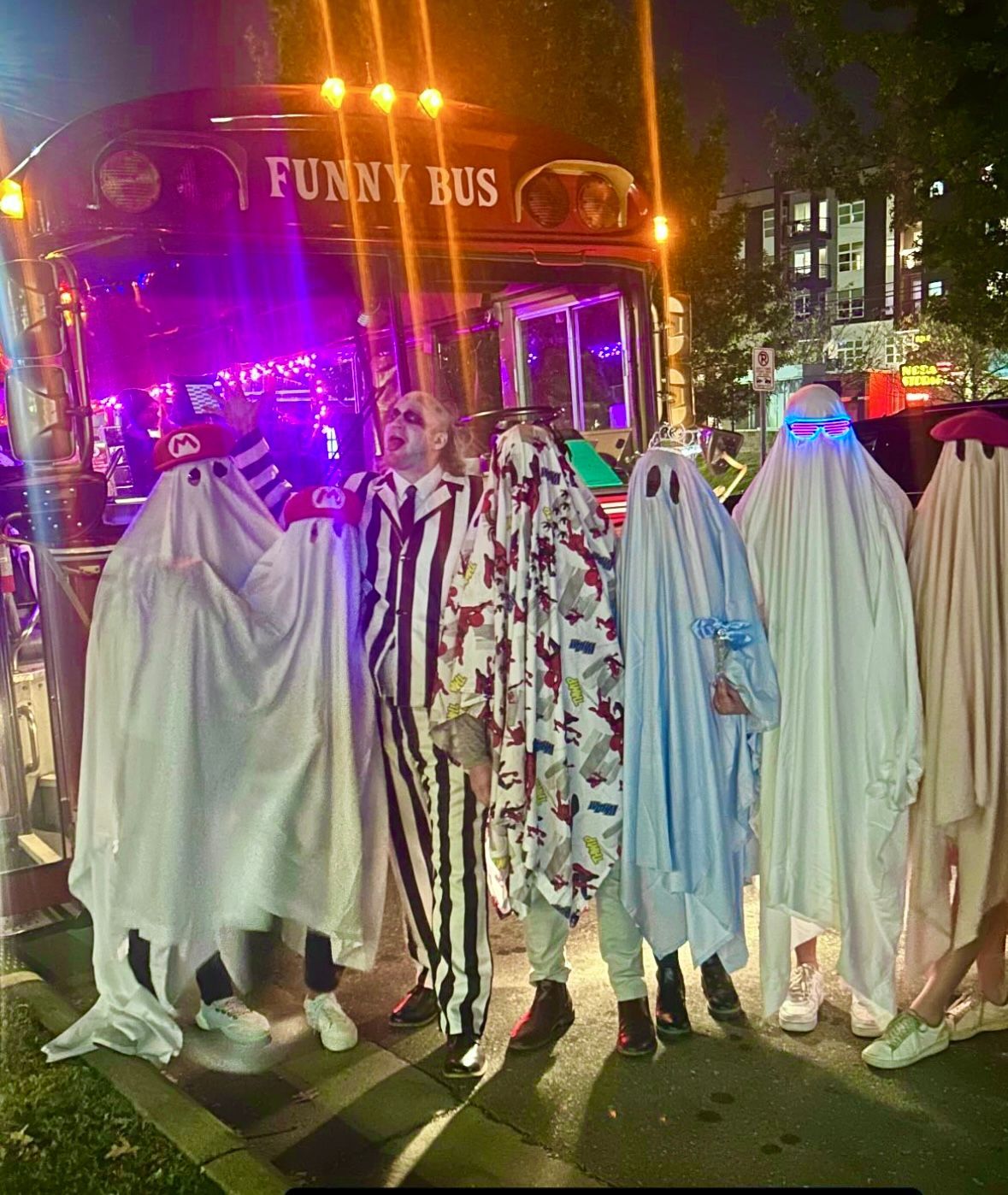 People in ghost costumes posing on a city street at night, with a lit bus behind them.