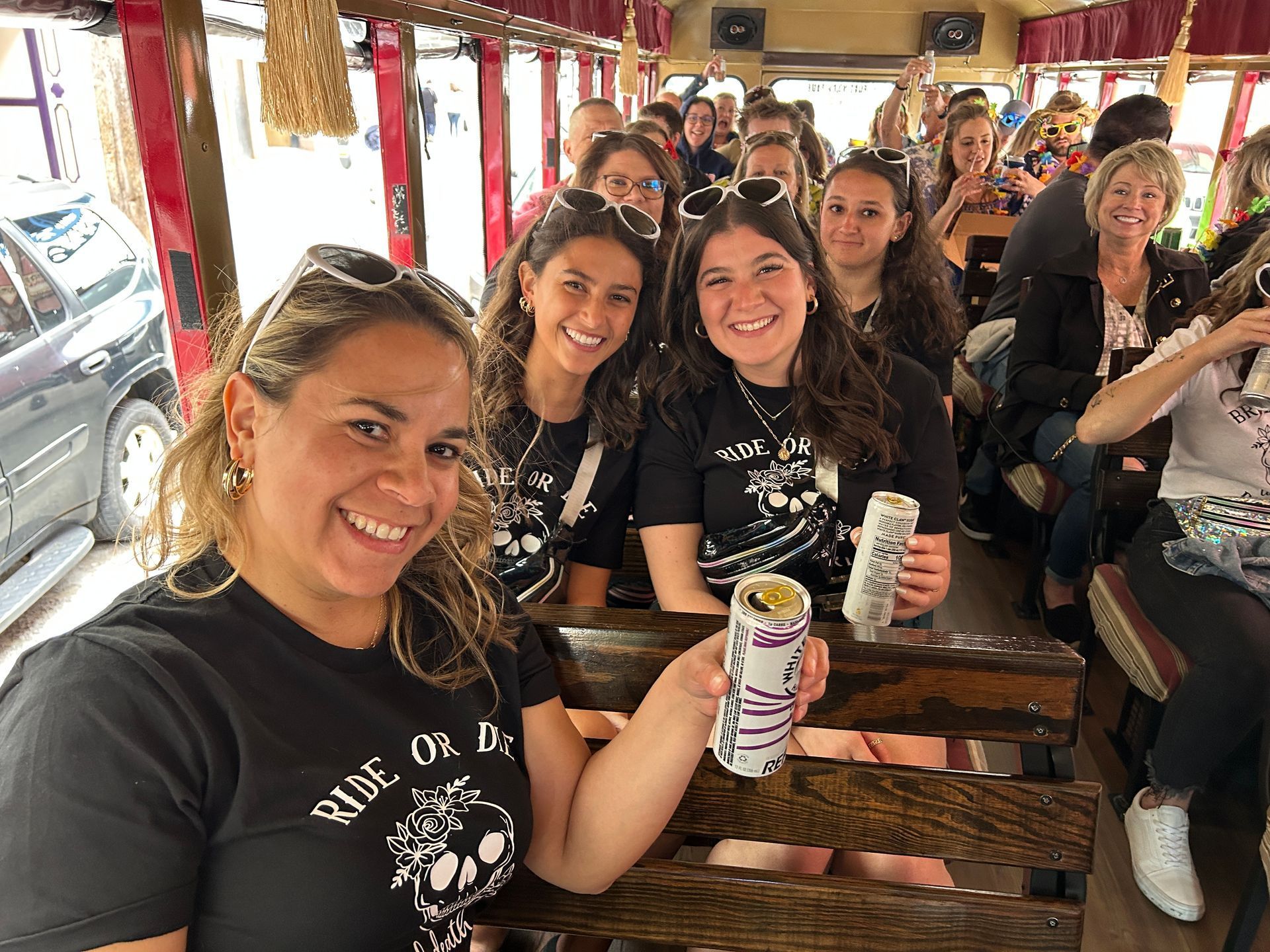 Smiling friends seated at a wooden table, holding drinks inside a crowded bus-like vehicle.