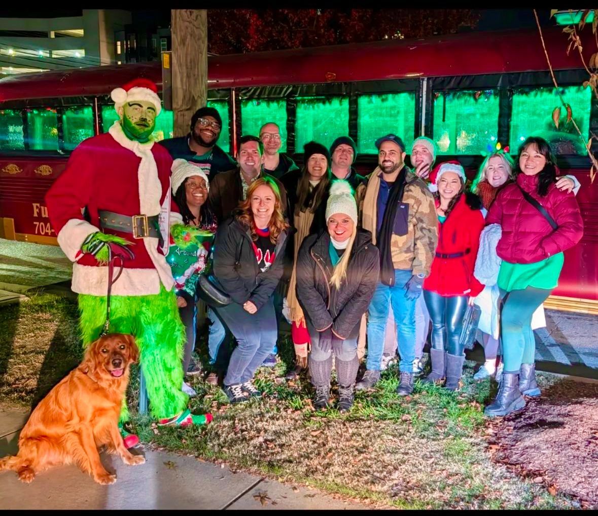 Group posing with Santa and a dog in front of a brightly lit holiday display at night