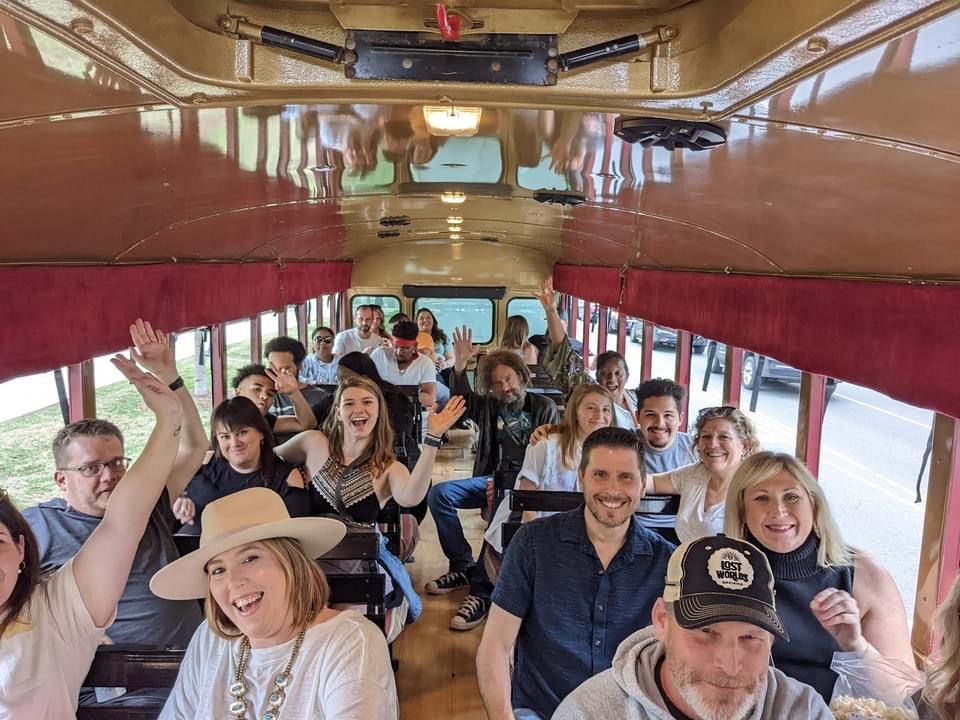 Group of smiling passengers riding in a vintage trolley bus with red curtains and wood-paneled interior.