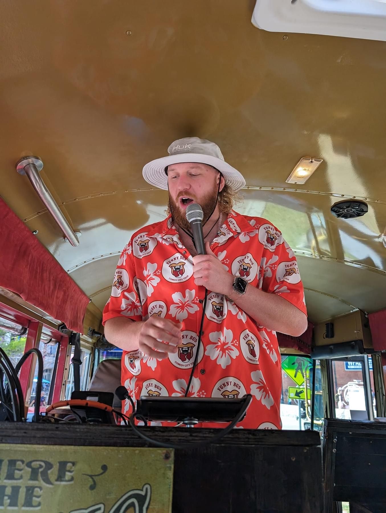 Man in red Hawaiian shirt and white hat speaking into a microphone on a bus stage
