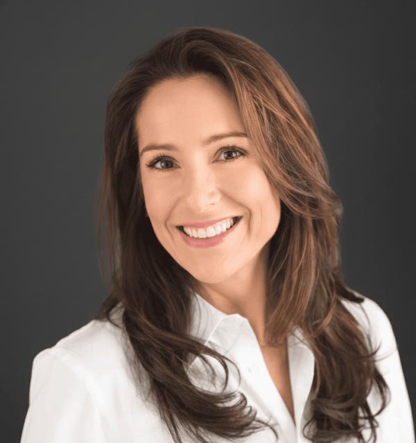 Woman with long brown hair, smiling, wearing a white shirt, against a dark background.