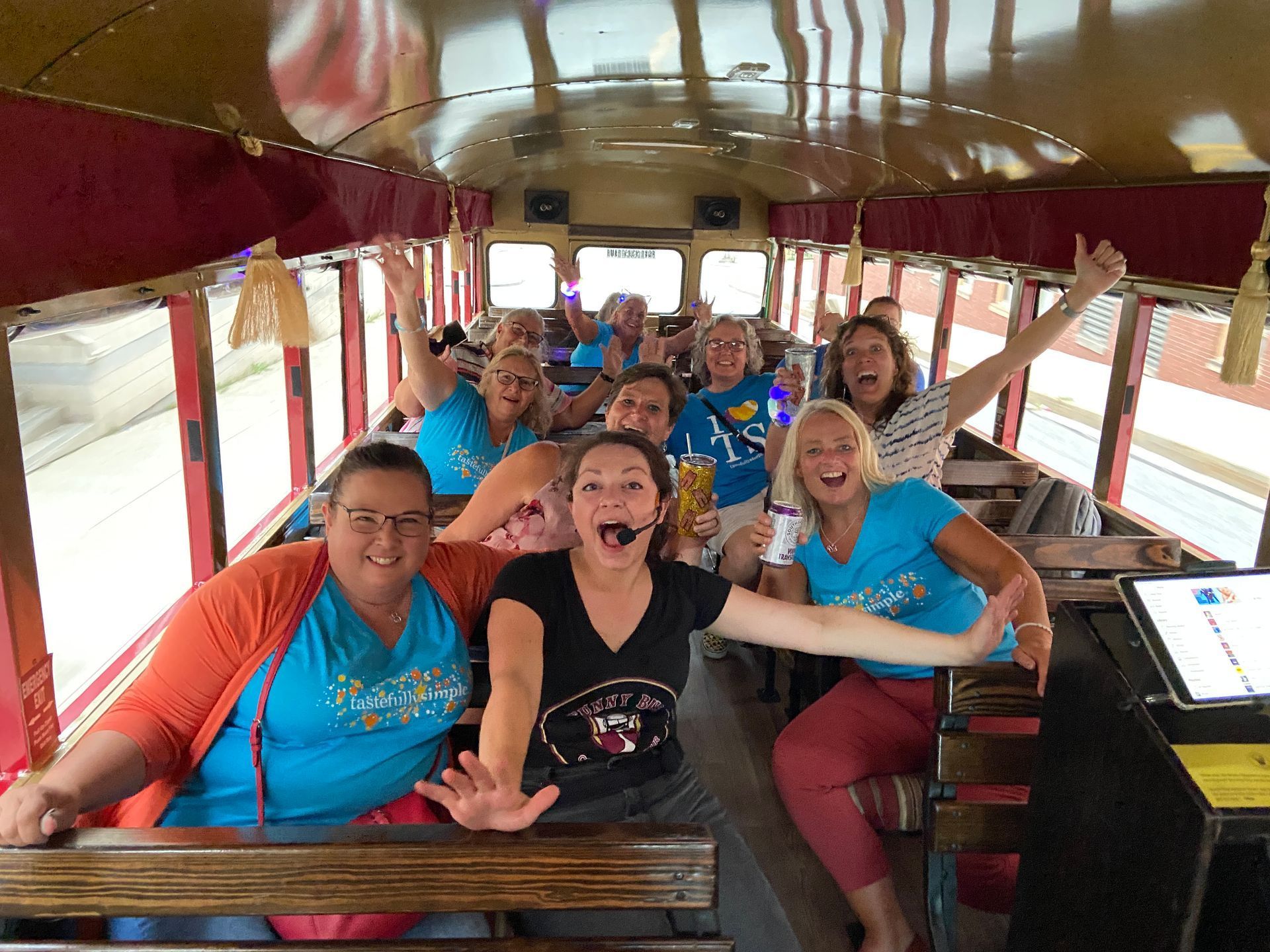 Group of smiling people seated on a trolley bus, waving inside a wood-paneled carriage.
