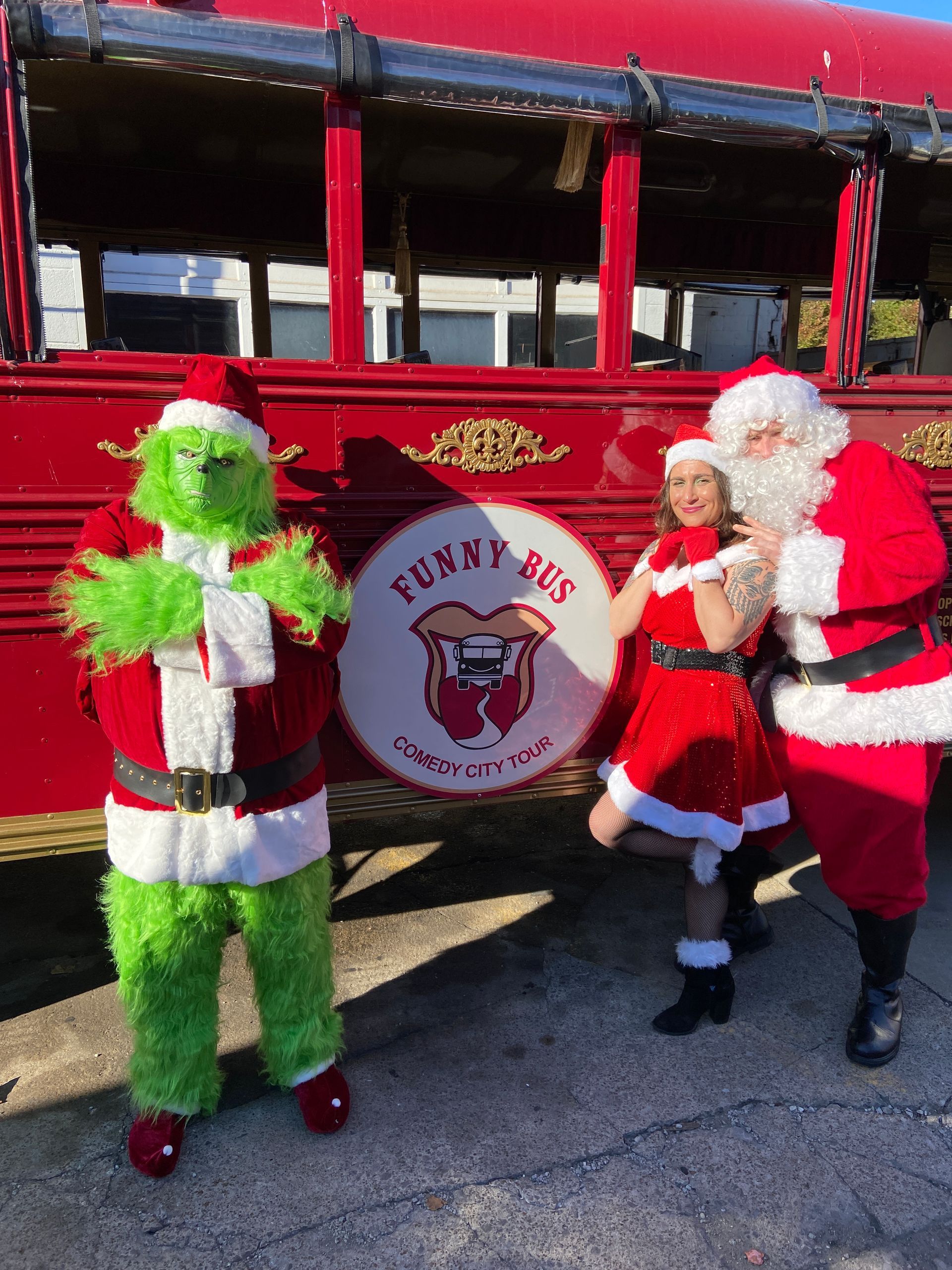 Santa and a green-costumed figure pose beside a red bus with a holiday logo.