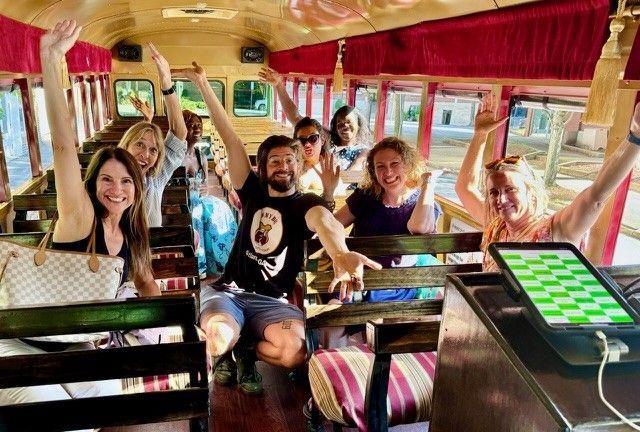 Passengers smiling and waving inside a colorful open-air vehicle with red curtains and wooden seats