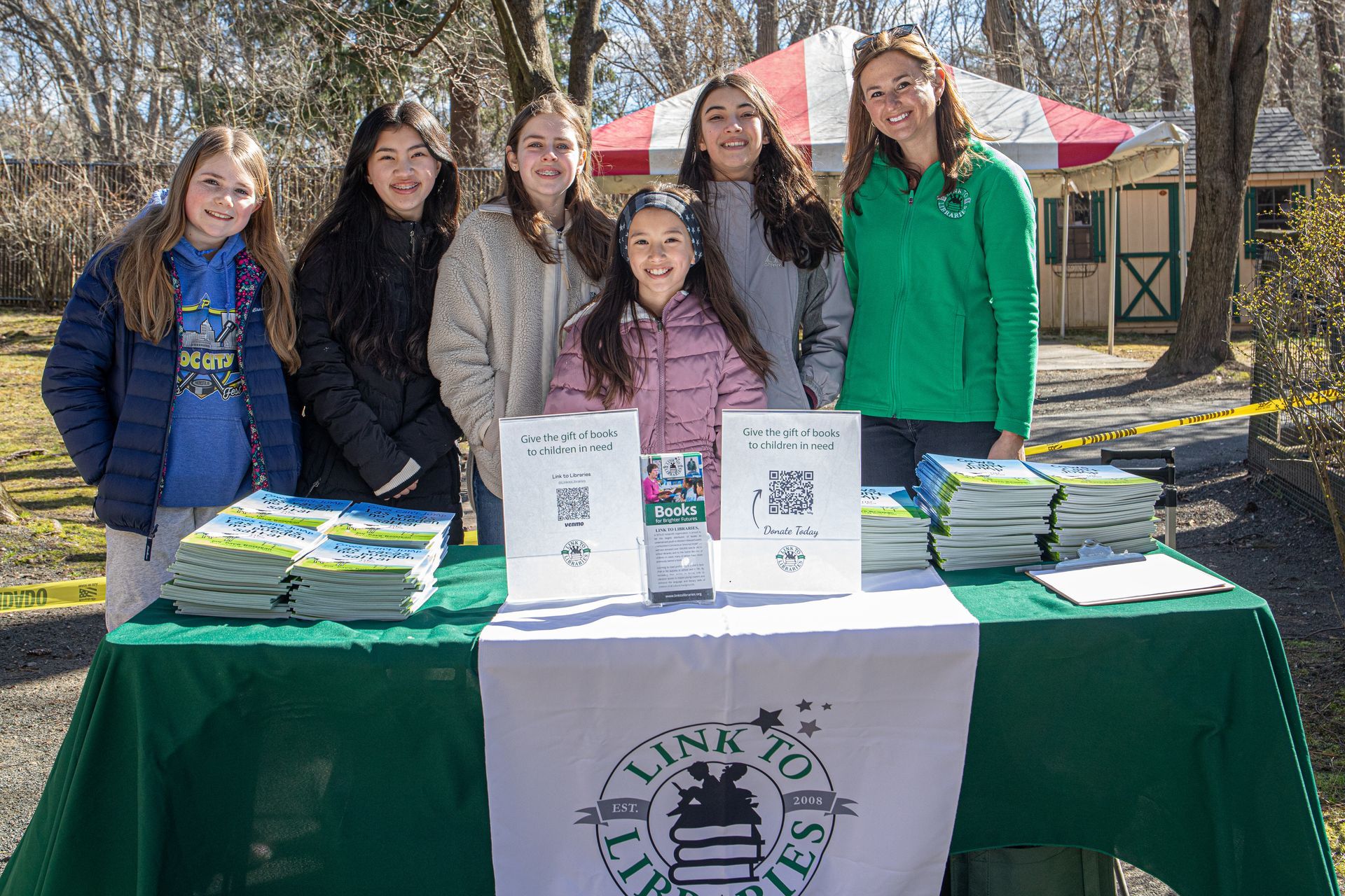 A group of volunteers associated with Link to Libraries stands behind a branded table covered in picture books.