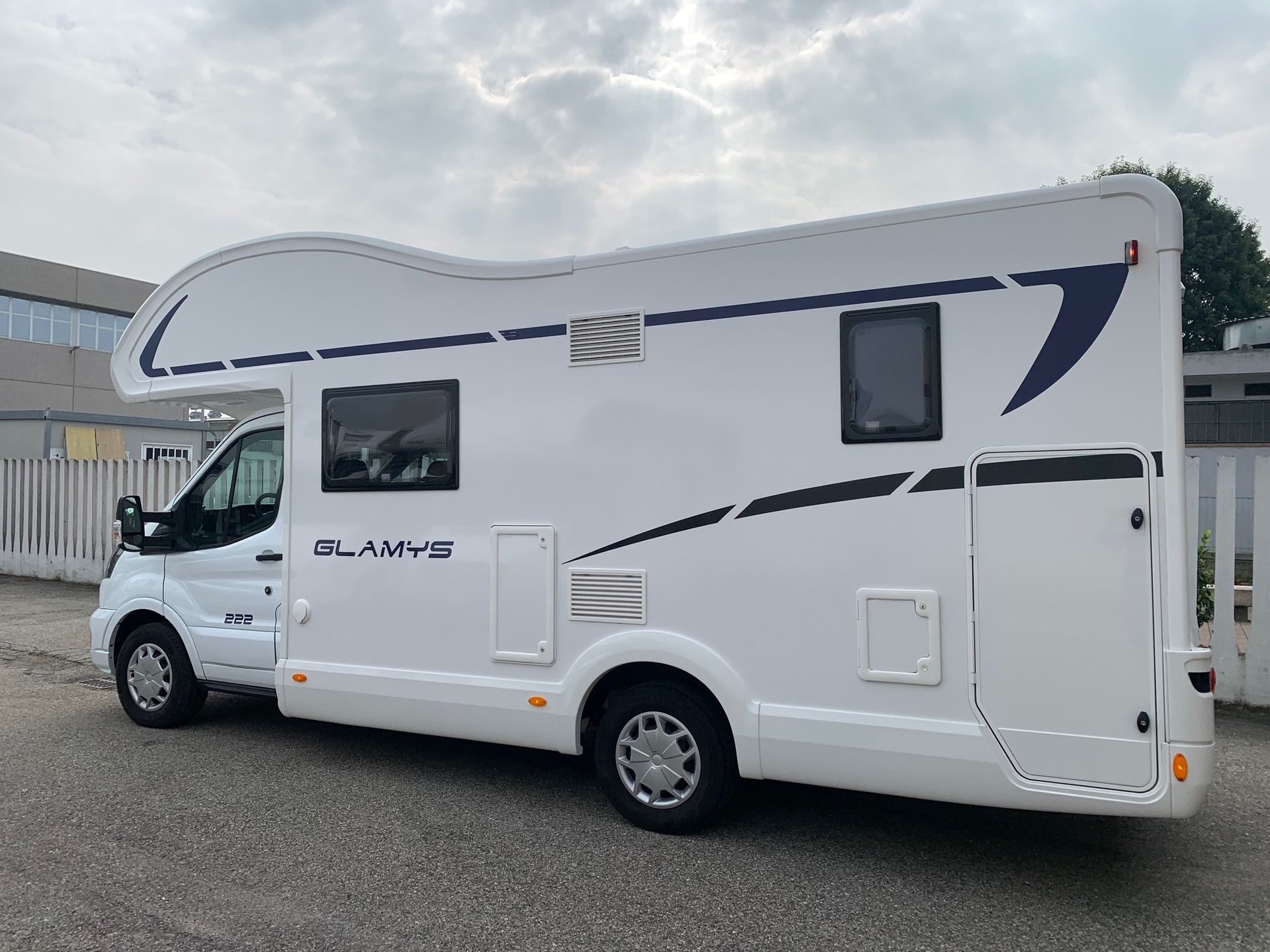 Camper bianco con decorazioni blu parcheggiato all'aperto sotto un cielo nuvoloso.