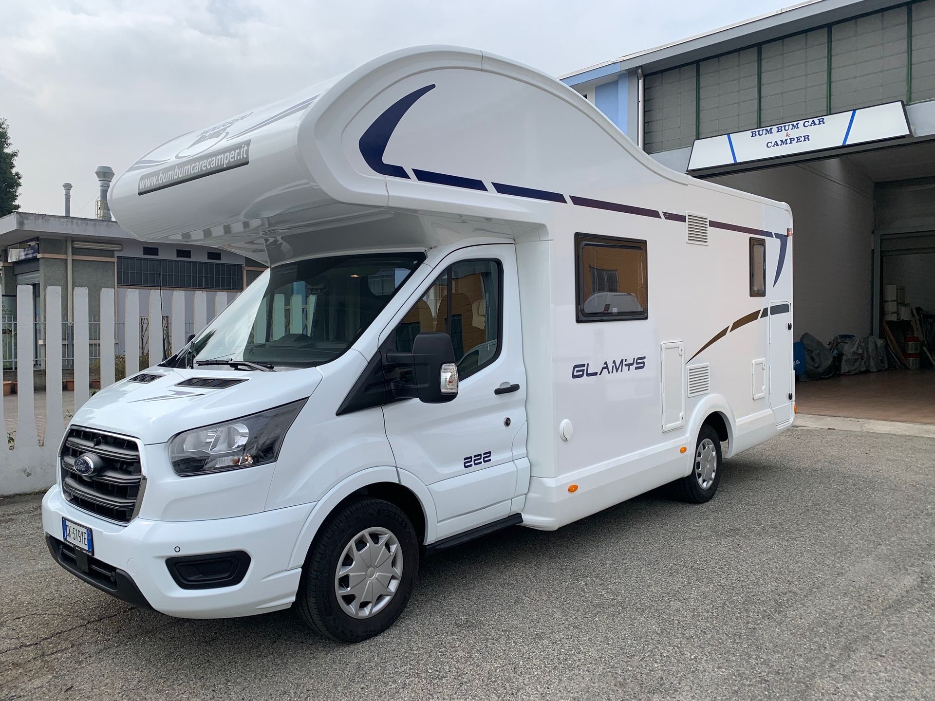 Un camper bianco parcheggiato fuori da un edificio, con una zona notte sopra la cabina e una striscia blu.