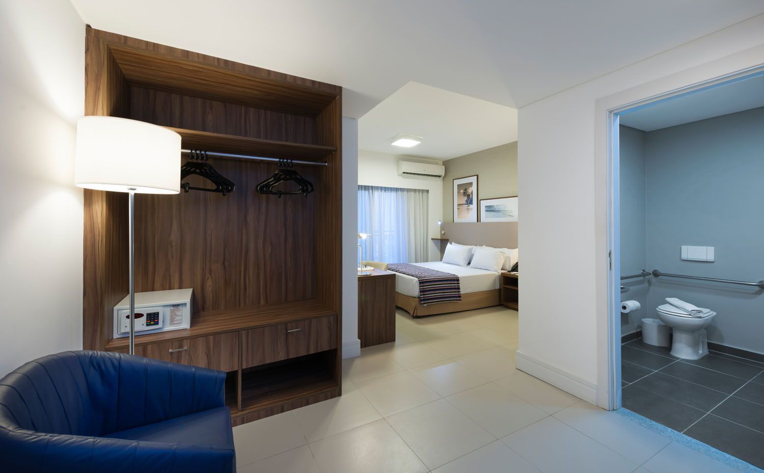 Um quarto de hotel com uma cama, cadeira, luminária e banheiro.