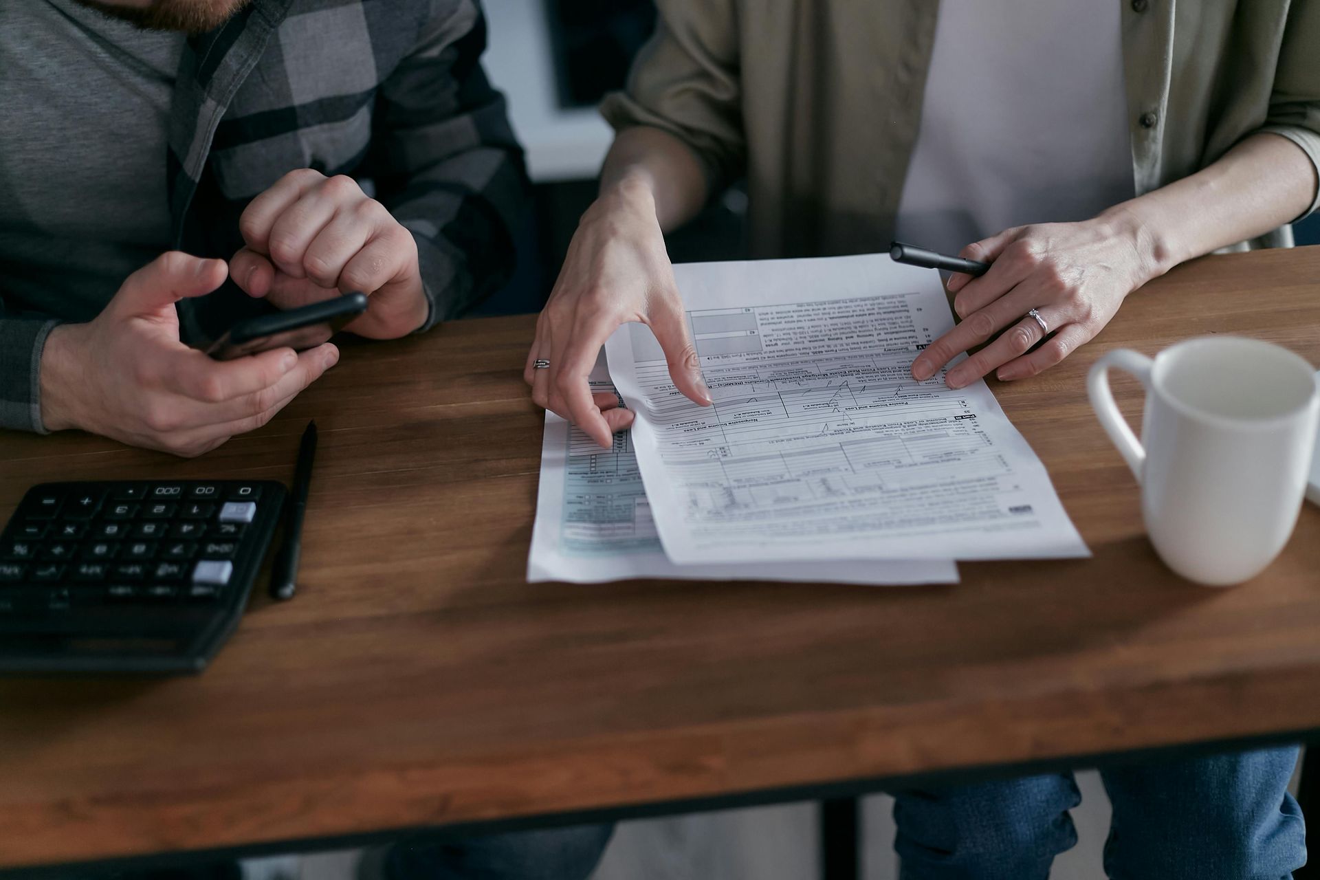 Couple reviewing financial documents with a calculator, phone, and mug on a wooden table.