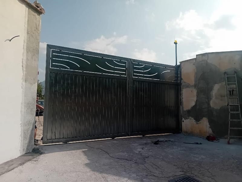 Un grande cancello di metallo grigio è parzialmente aperto. Si trova di fronte a un edificio di cemento e al cielo.