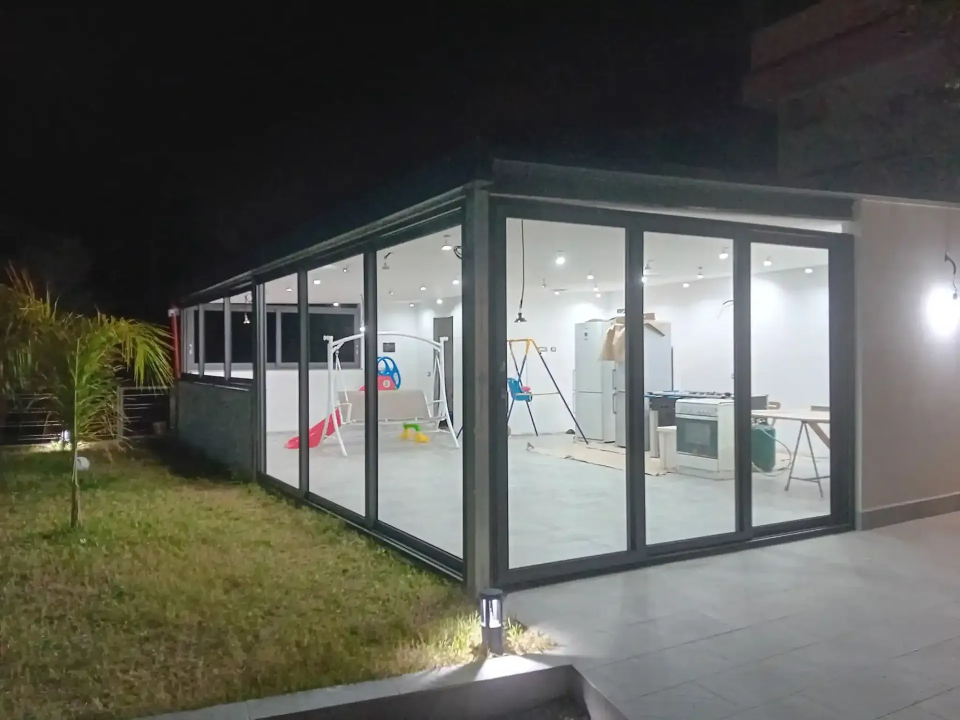 Stanza con pareti in vetro, luci interne accese, mobili e oggetti a vista. All'esterno, prato e qualche luce accesa di notte.